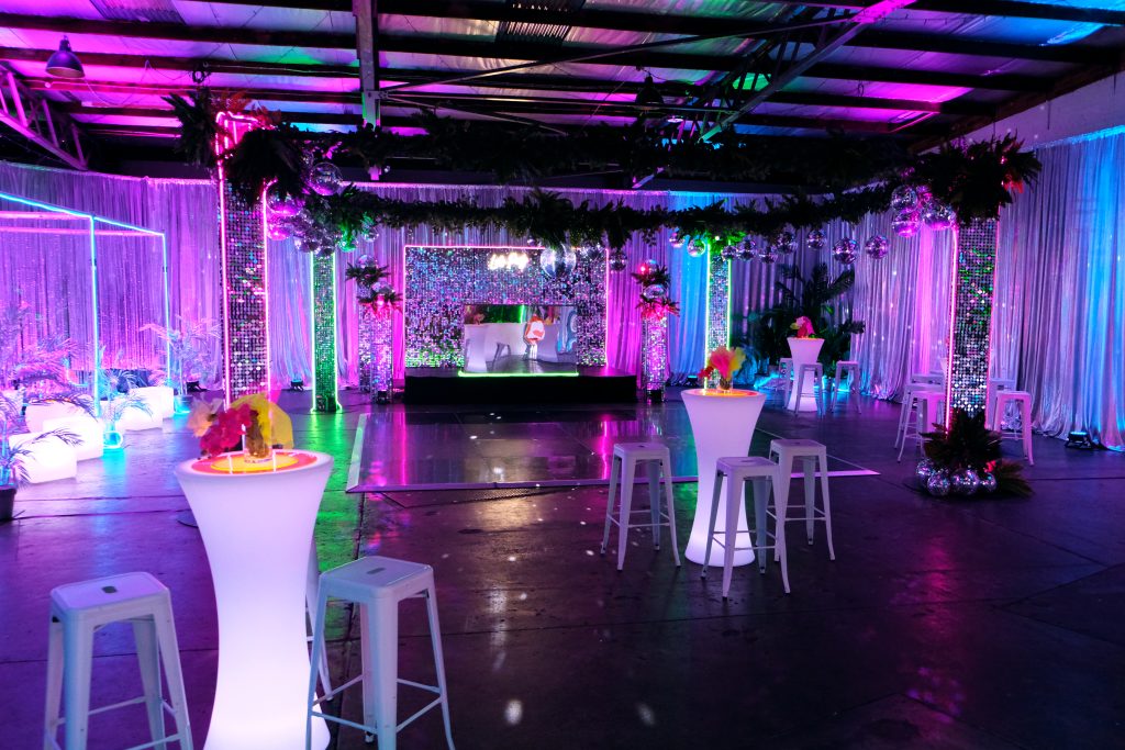 Room shot of neon disco party theme featuring illuminated furniture, bar stools, greenery, dance floor, neon lights, and sequin panels neon disco theme decorating essentials