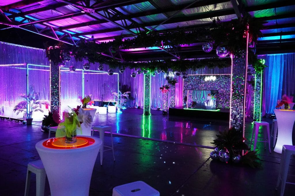 Room shot of neon disco party theme featuring illuminated furniture, bar stools, greenery, dance floor, neon lights, and sequin panels how to pick the right party theme