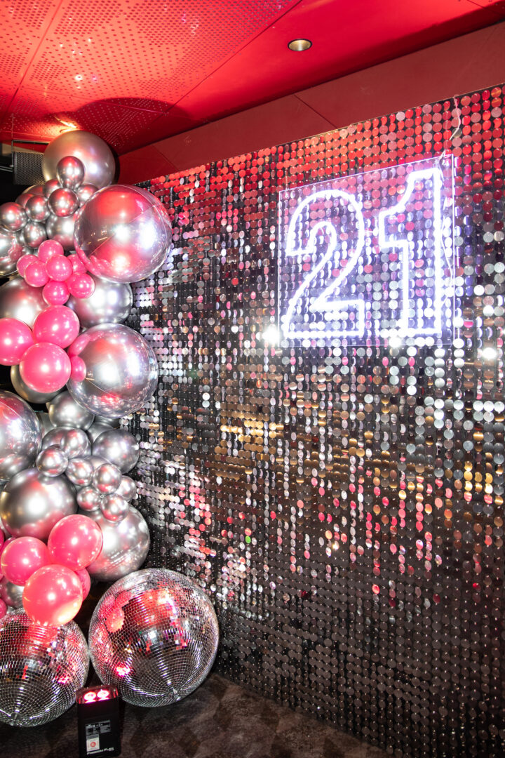 Silver sequin birthday backdrop with silver and pink balloon garland, 21 neon sign, and mirror balls