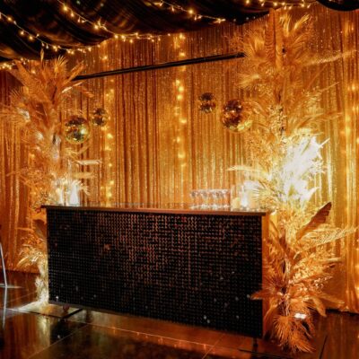 bar area with gold sequin drape, gold leaves, gold mirror balls, black roof drape, and fairy lights at black and gold themed setup