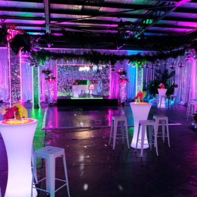 Room shot of neon disco party theme featuring illuminated furniture, bar stools, greenery, dance floor, neon lights, and sequin panels neon disco theme decorating essentials