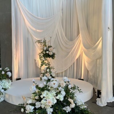 2025 wedding décor trends Gathered dramatic drape wedding ceremony backdrop with white stage and lush white and green florals